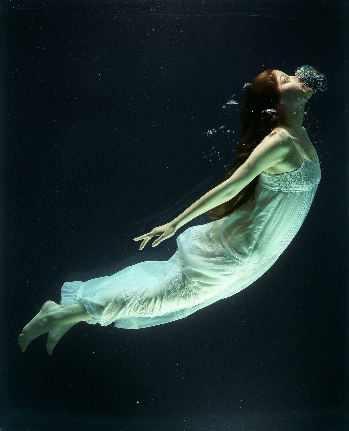 Eine Frau in einem weißen Kleid unter Wasser mit Luftblasen um Mund und Nase